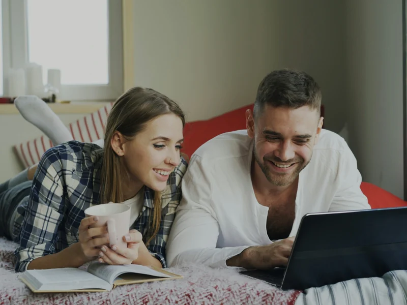 Ein Paar liegt entspannt mit Laptop und Kaffee auf dem Bett